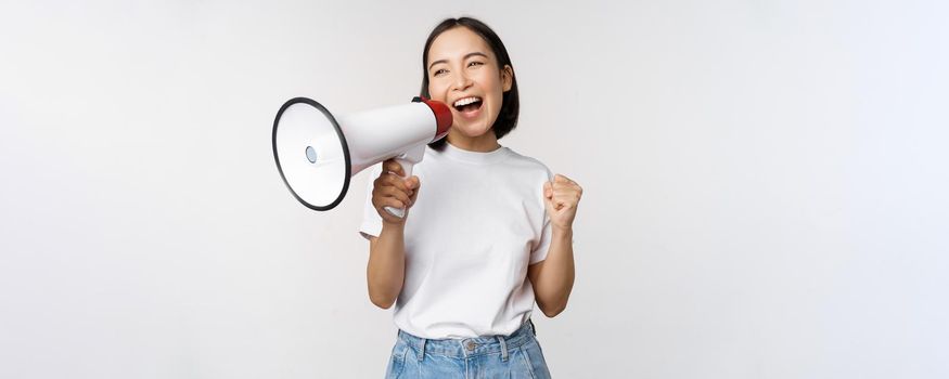 Asian girl shouting at megaphone, young activist protesting, using loud speakerphone, making announcement, white background. Copy space