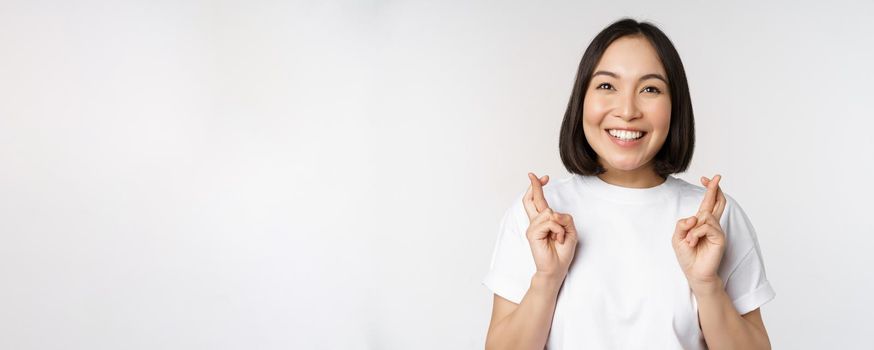 Hopeful asian girl cross fingers, making wish, anticipating, wishing smth, standing in tshirt over white background. Copy space