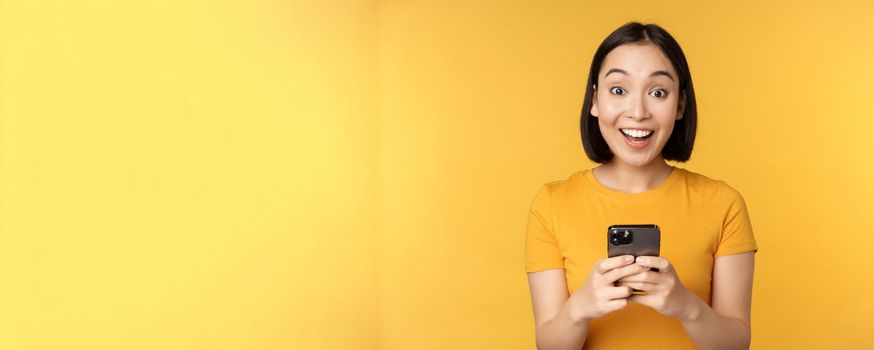 Happy asian girl smiling, standing with black mobile phone, standing against yellow background. Copy space