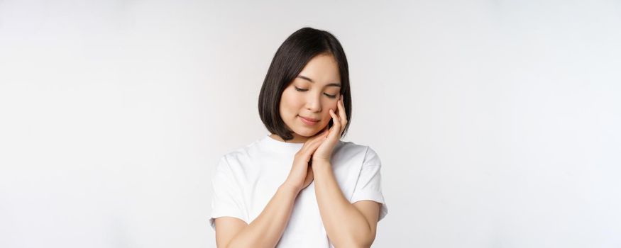 Portrait of tender and feminine young asian woman, looking down, gently touching face, concept of skin care and cosmetology, white background.