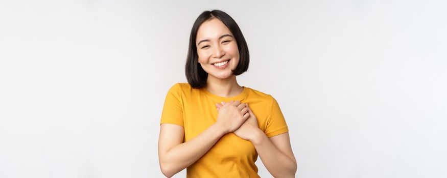 Romantic asian girfriend, holding hands on heart, smiling with care and tenderness, standing in yellow tshirt over white background.