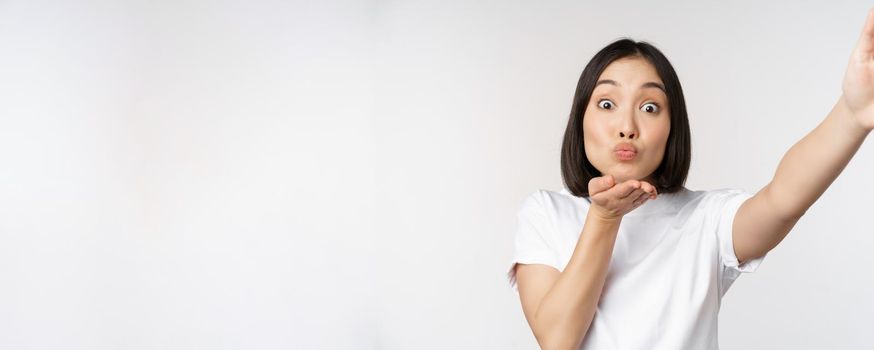 Beautiful young asian woman smiling, looking at camera, holding device, taking selfie, video chat, standing in tshirt over white background.