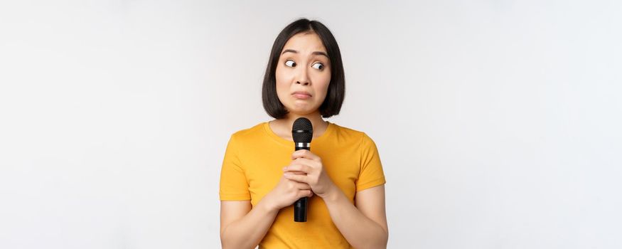 Modest asian girl holding microphone, scared talking in public, standing against white background. Copy space