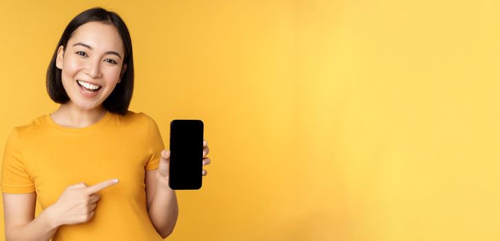 Beautiful happy asian girl showing mobile phone screen, application on smartphone gadget, standing over yellow background.