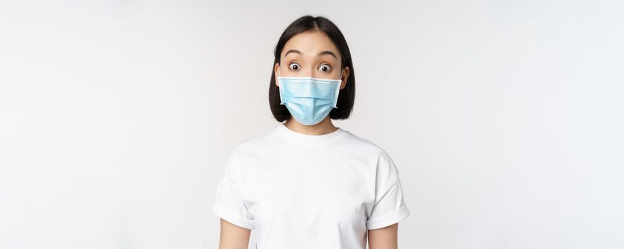 Health and covid pandemic concept. Image of korean woman in face medical mask looking surprised, reacting amazed, standing over white background.