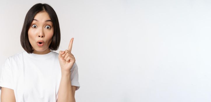 Close up of creative asian girl raising finger, suggesting smth, has an idea, pointing up, eureka sign, standing over white background.