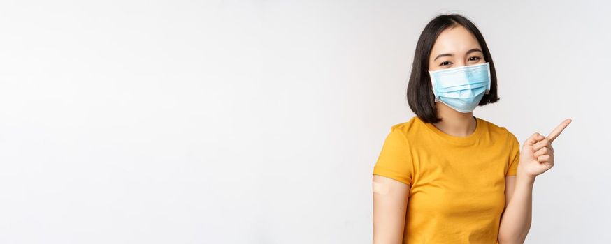 Covid-19, vaccination and healthcare concept. Portrait of cute asian girl in medical mask, has band aid on shoulder after coronavirus vaccine, standing over white background.