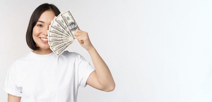 Portrait of smiling asian woman holding dollars money, concept of microcredit, finance and cash, standing over white background.