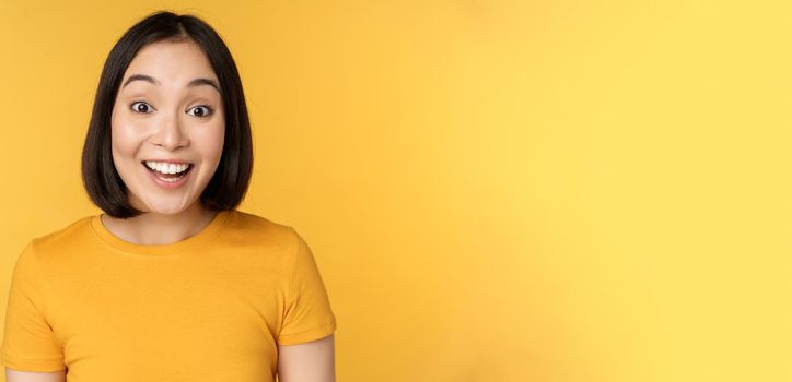 Close up portrait of asian girl showing surprised reaction, raising eyebrows amazed, reacting to big news, standing over yellow background.