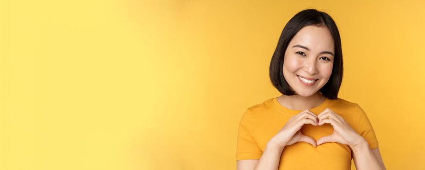 Close up portrait of smiling korean woman, showing romantic heart sign and looking happy, standing over yellow background. Copy space