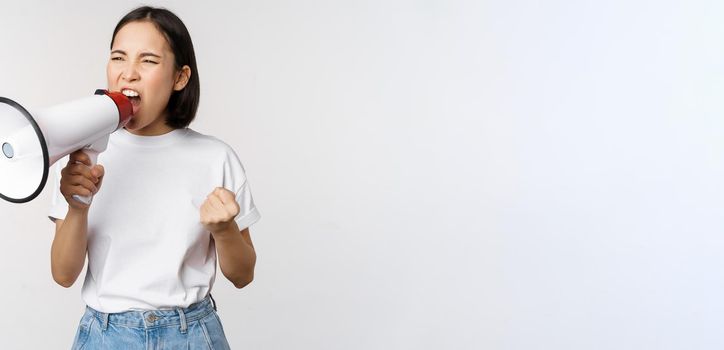 Confident asian woman shouting in megaphone, screaming and protesting. Girl activist using speaker to speak louder, standing over white background.