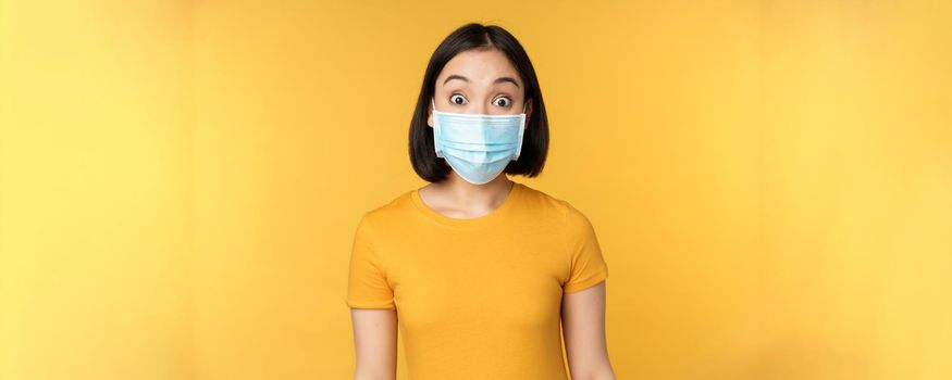 Portrait of korean girl in medical face mask looking surprised, amazed reaction to news, standing over yellow background.