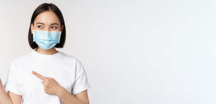 Young korean woman in medical face mask pointing fingers left and looking at logo, showing advertisement or banner, standing over white background.