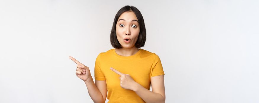 Portrait of smiling asian brunette girl in yellow tshirt, pointing fingers left, showing copy space, promo deal, demonstrating banner, standing over white background.