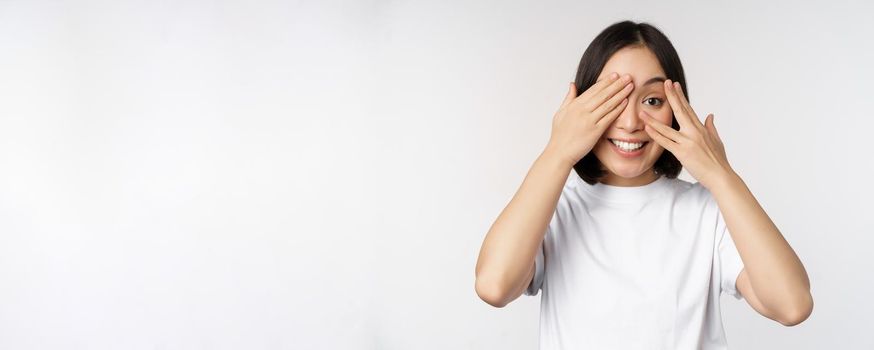 Portrait of asian woman covering eyes, waiting for surprise blindfolded, smiling and peeking at camera, standing over white background.