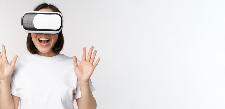 Happy asian woman using VR headset, waving raised hands and laughing, using virtual reality glasses, standing over white background.