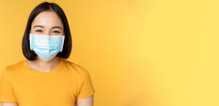 Close up of smiling happy asian woman, wearing medical face mask from covid-19, standing in yellow t-shirt over studio background.