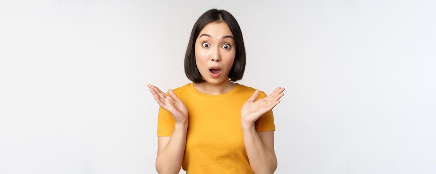 Close up portrait of asian woman looking surprised, wow face, staring impressed at camera, standing over white background in yellow t-shirt.