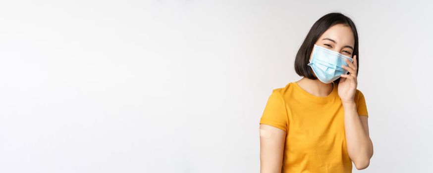 Covid-19, vaccination and healthcare concept. Cute asian girl in medical face mask, showing band aid after coronavirus vaccination, standing over white background.