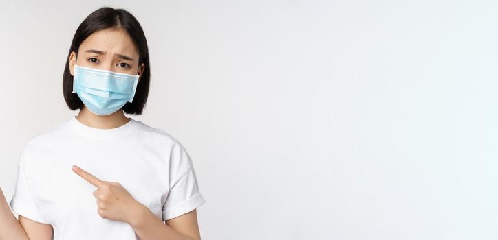 Sad asian woman in medical mask, pointing fingers left, frowning and looking upset, complaining, demonstrating banner, standing over white background.