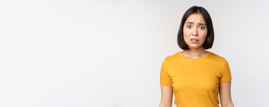 Portrait of worried korean girl, looking concerned at camera, standing in yellow tshirt over white background.