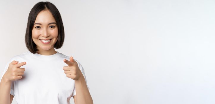 Image of smiling asian girl pointing fingers at camera, choosing, inviting you, congratulating, standing in tshirt over white background.