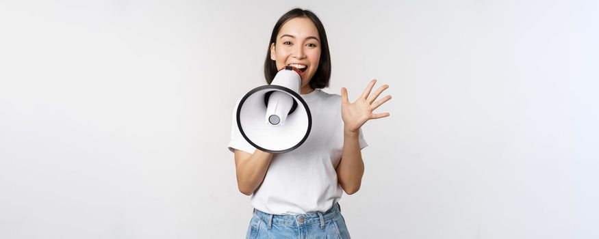 Happy asian woman shouting at megaphone, making announcement, advertising something, standing over white background.