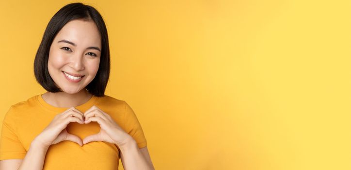Close up portrait of smiling korean woman, showing romantic heart sign and looking happy, standing over yellow background. Copy space