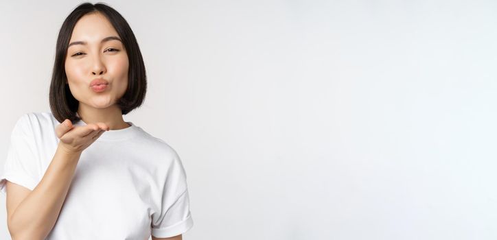 Lovely young asian woman, sending air kiss and looking coquettish at camera, standing in tshirt over white background.