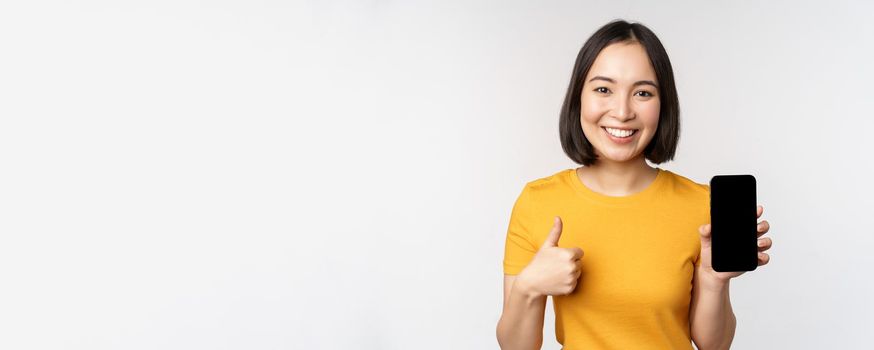 Technology and people concept. Smiling young woman showing thumb up and smartphone screen, mobile phone app interface, standing against white background.