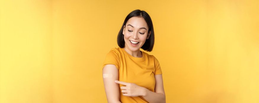 Vaccination and covid-19 pandemic concept. Happy and healthy asian girl pointing at her shoulder with band aid after vaccinating from coronavirus, yellow background.