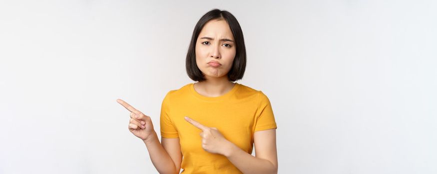 Portrait of disappointed brunette asian girl, pointing fingers left, grimacing upset, showing smth unpleasant, standing over white background.