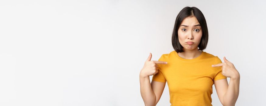 Confused asian woman pointing at herself, looking in disbelief, standing over white background. Copy space