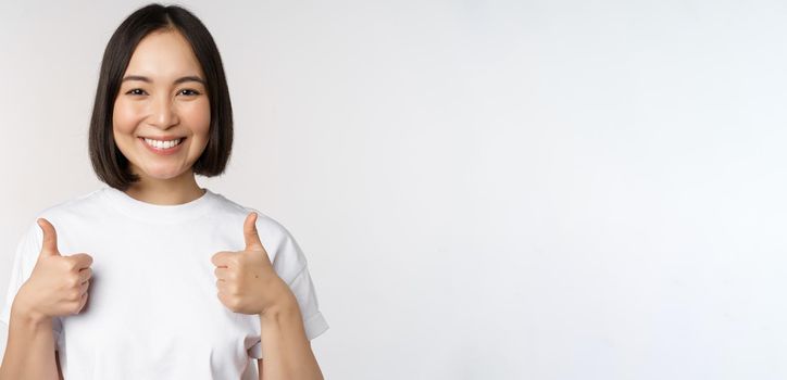 Happy young korean woman showing thumbs up and smiling, give positive feedback, recommending smth, standing over white background.