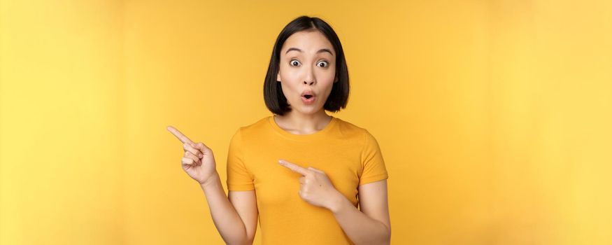 Smiling asian woman pointing fingers left, showing advertisement on empty copy space, standing over yellow background.