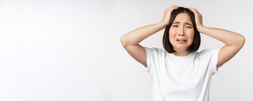 Desperate young korean woman holding hands on head, panicking, crying and standing distressed against white background.