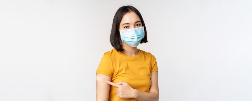 Covid-19, vaccination and healthcare concept. Portrait of cute asian girl in medical mask, has band aid on shoulder after coronavirus vaccine, standing over white background.