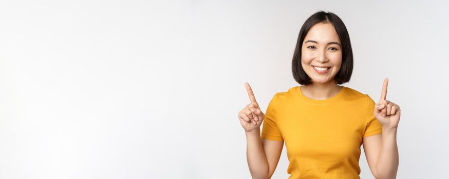 Portrait of beautiful japanese girl smiling, pointing fingers up, showing advertisement, standing in yellow tshirt against white background.
