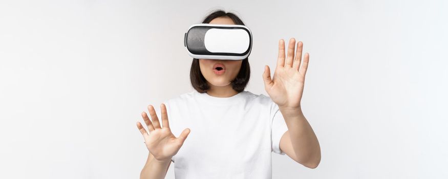 Amused asian girl using VR glasses, virtual reality headset and reaching hands into empty space, touching smth augmented, standing over white background.