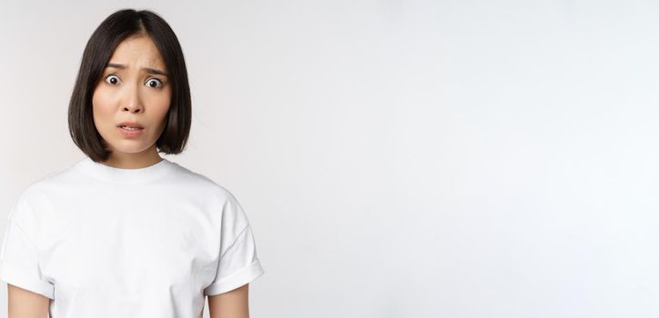 Image of shocked and frustrated asian woman, looking alarmed at camera, cant understand, puzzled, standing over white background.