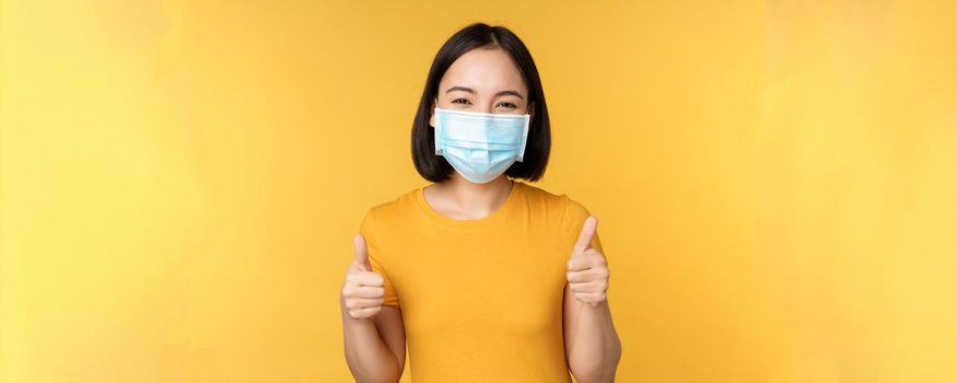 Smiling asian woman in medical face mask, showing thumbs up approval, like and recommend smth, standing over yellow background.