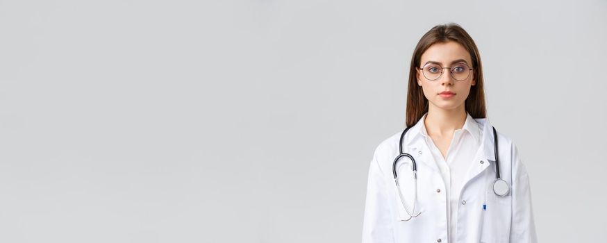 Healthcare workers, medicine, insurance and covid-19 pandemic concept. Serious-looking smart professional doctor, nurse in white scrubs and stethoscope, look camera determined, grey background.