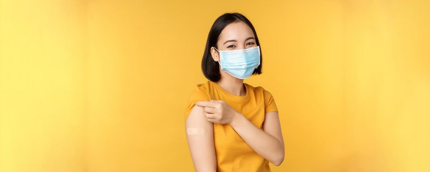 Vaccination and covid-19 pandemic concept. Smiling asian woman in medical face mask, showing her shoulder with band aid after vaccinating from coronavirus, yellow background.