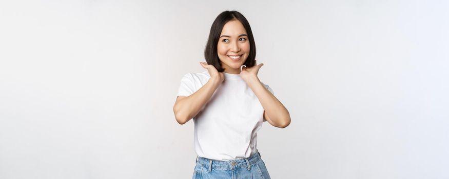 Portrait of cute, beautiful asian woman touching her new short haircut, showing hairstyle, smiling happy at camera, standing over white background.