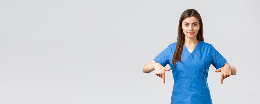 Healthcare workers, prevent virus, insurance and medicine concept. Smiling pretty nurse or doctor in blue scrubs, pointing fingers down, showing patients advertisement, recommend drugstore.