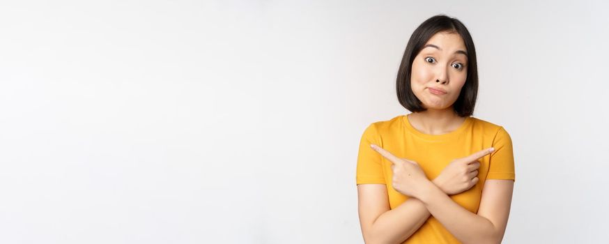 Complicated asian girl pointing fingers sideways, showing left and right choices, staring indecisive clueless, standing over white background. Copy space