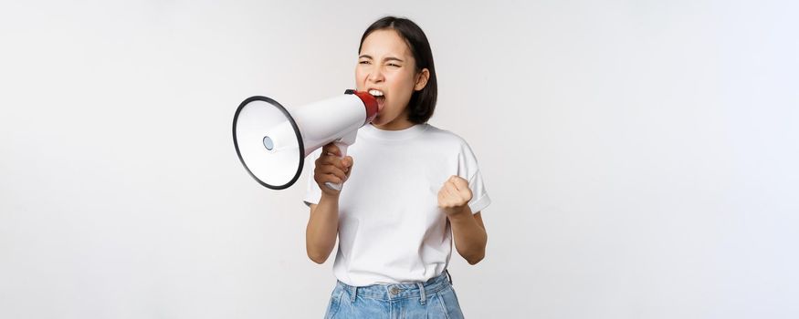 Confident asian woman shouting in megaphone, screaming and protesting. Girl activist using speaker to speak louder, standing over white background.