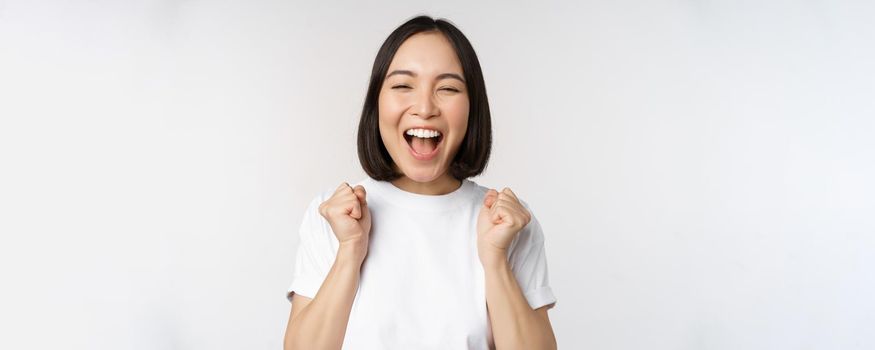 Portrait of enthusiastic asian woman winning, celebrating and triumphing, raising hands up, achieve goal or success, standing over white background.