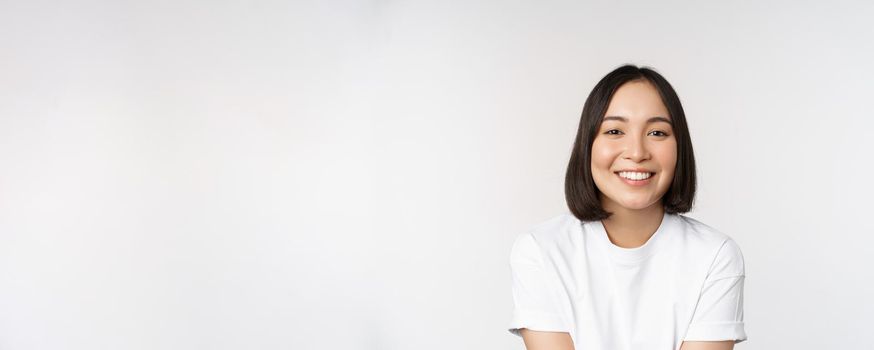 Beautiful korean girl smiling, white teeth, looking lovely at camera, standing in white tshirt over studio background.