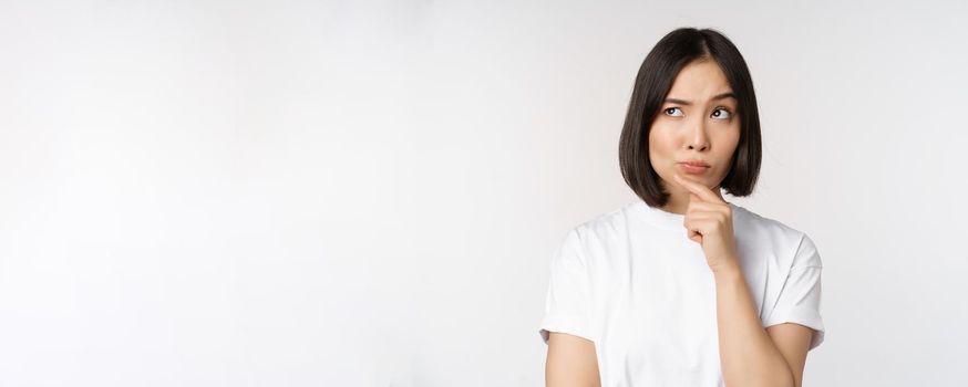 Thoughtful asian woman looking aside, pondering, making assumption, thinking or choosing smth, standing over white background.